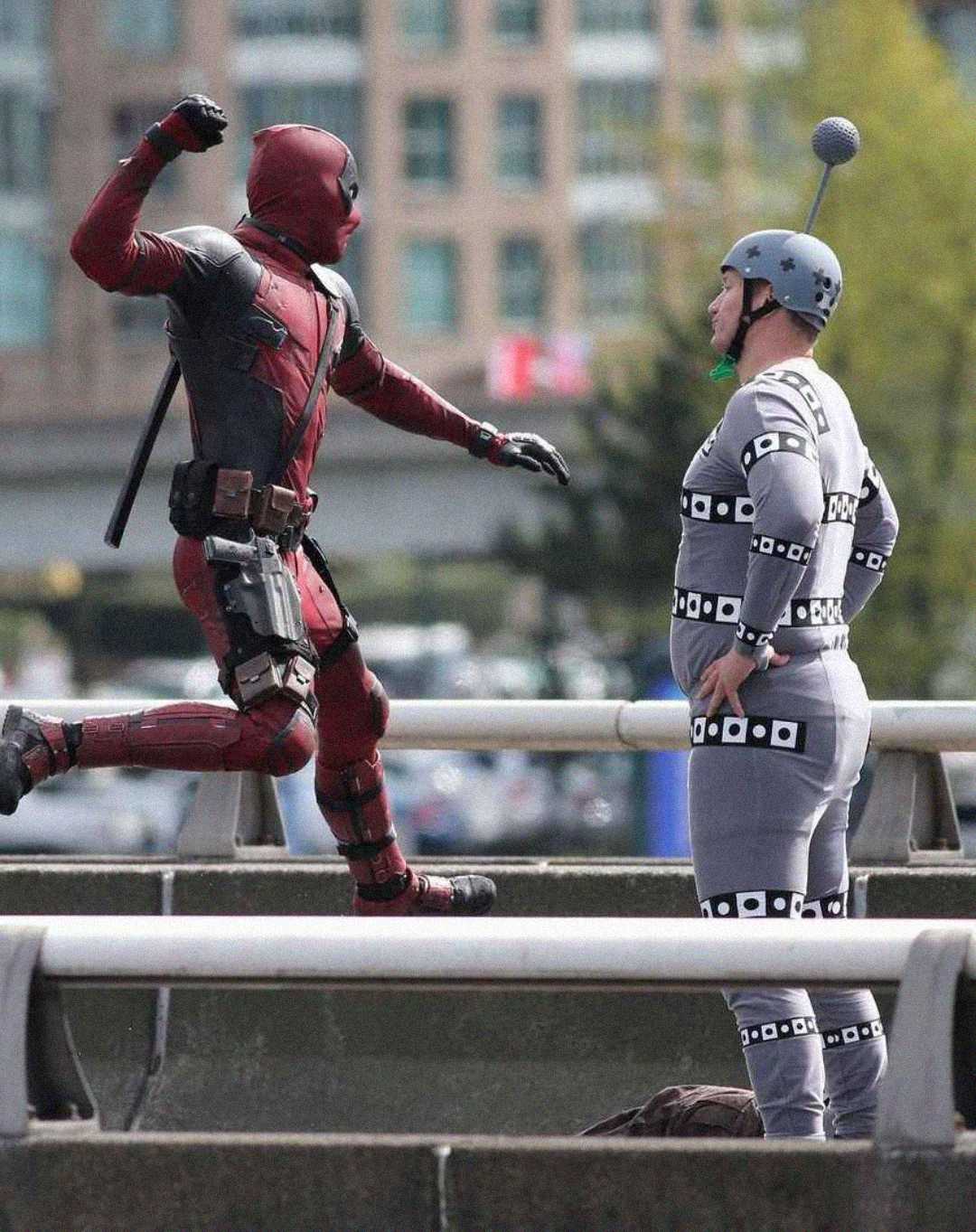A person in a Deadpool costume poses as if punching another person wearing a gray motion capture suit with tracking markers, standing on an outdoor bridge with buildings in the background.