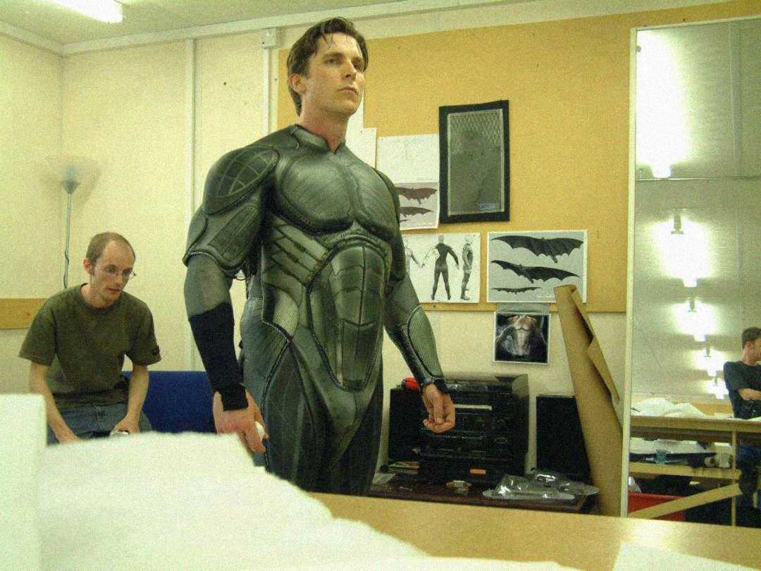 A man stands in a room wearing a detailed, futuristic black and grey suit, resembling superhero armor. Behind him, another man sits on a bench. The walls display sketches of bats and costume designs.