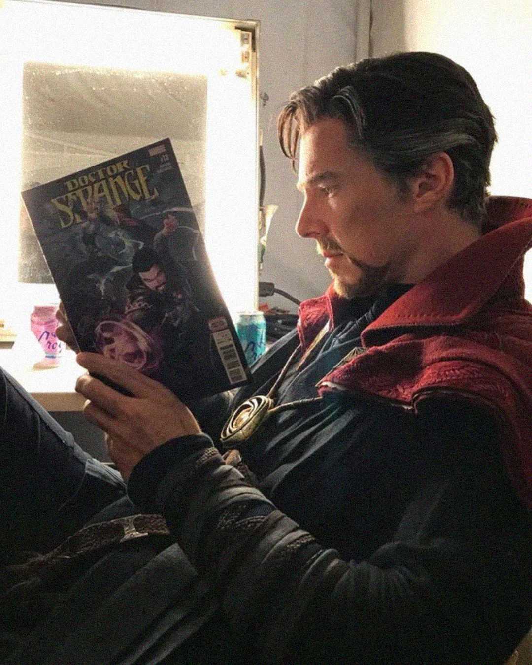 A man dressed as Doctor Strange sits by a window, intently reading a Doctor Strange comic book. He wears a red cloak and dark costume, with a focused expression on his face.