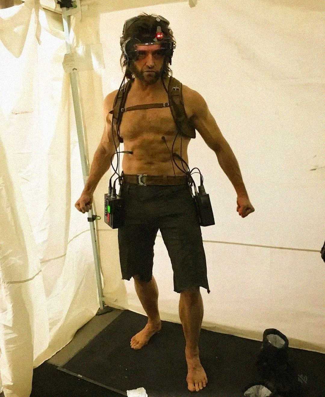 A barefoot, shirtless man stands in a tent, wearing black shorts and a headgear with wires and sensors attached to his chest, arms, and waist, striking a strong pose.