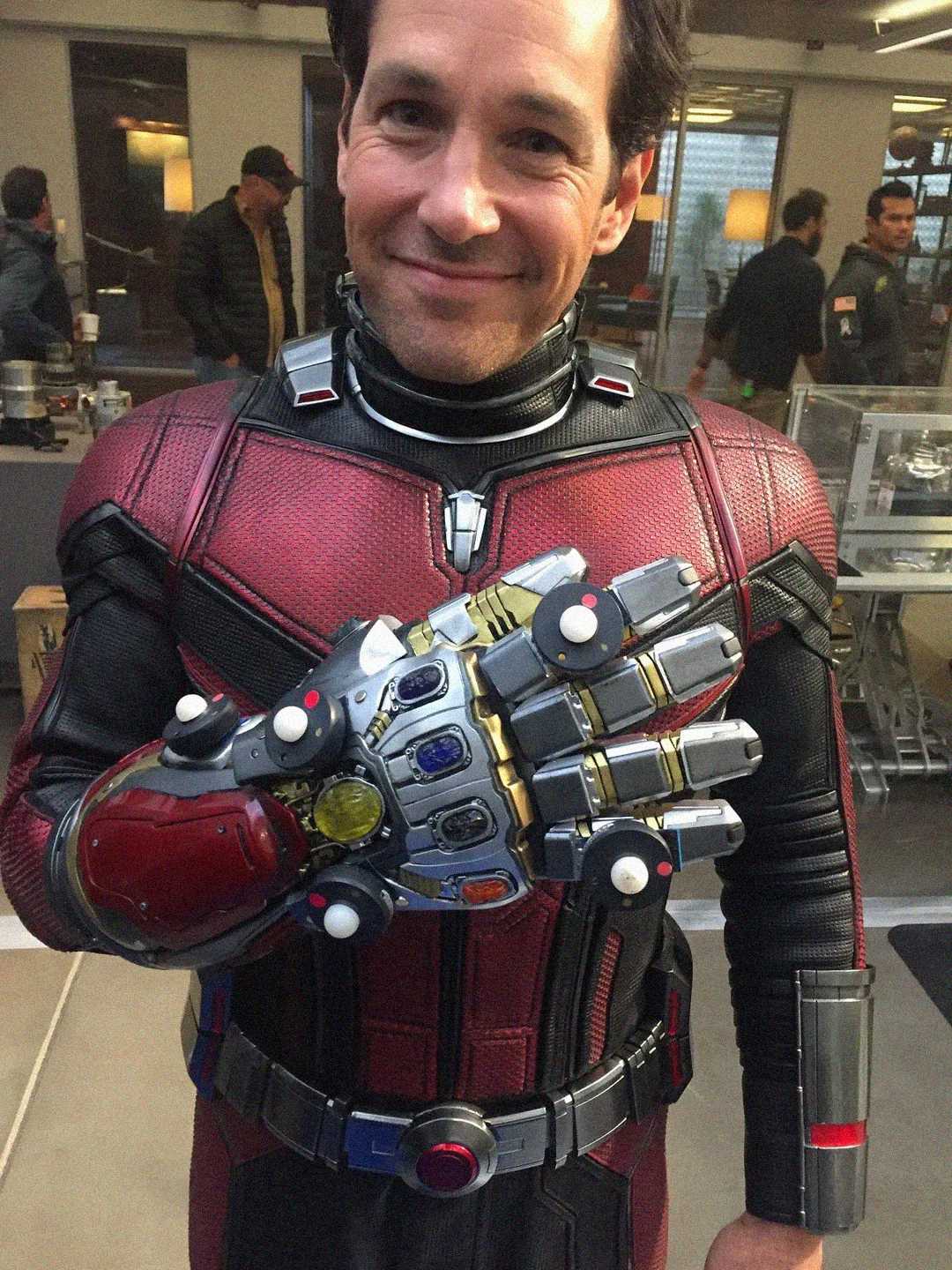 A man in a red and black superhero costume smiles at the camera while wearing a metallic glove with colorful stones, resembling the Infinity Gauntlet from the Marvel universe.