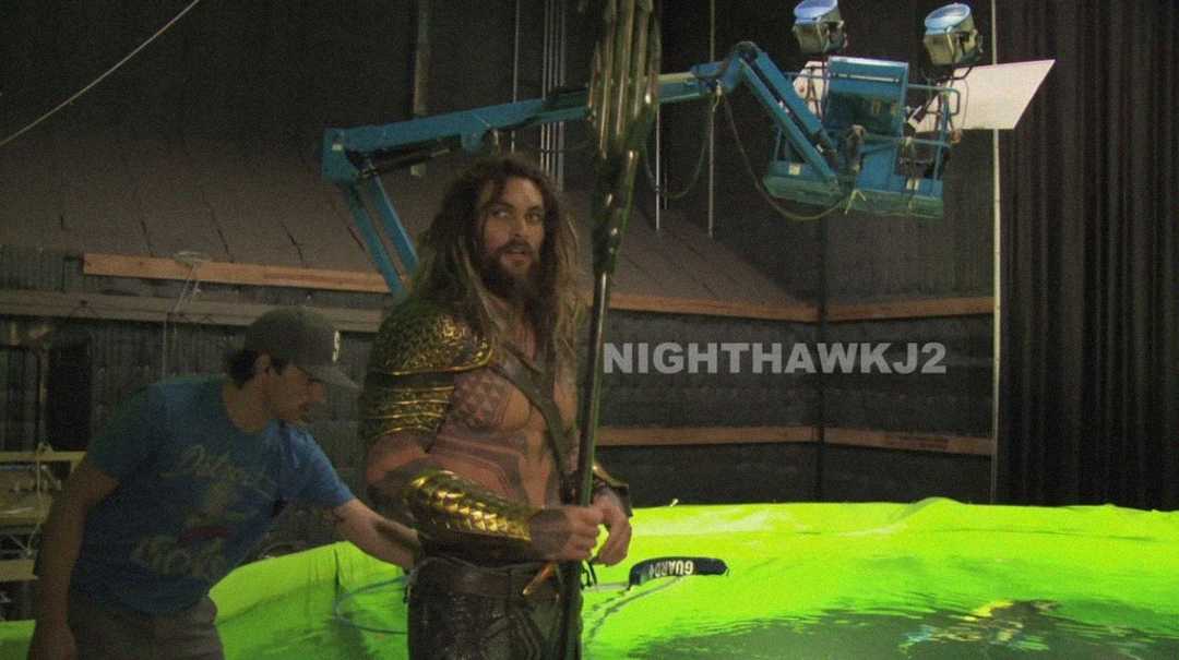 An actor in an Aquaman costume holds a trident on a film set, standing next to a green water tank with crew members and film equipment visible in the background.