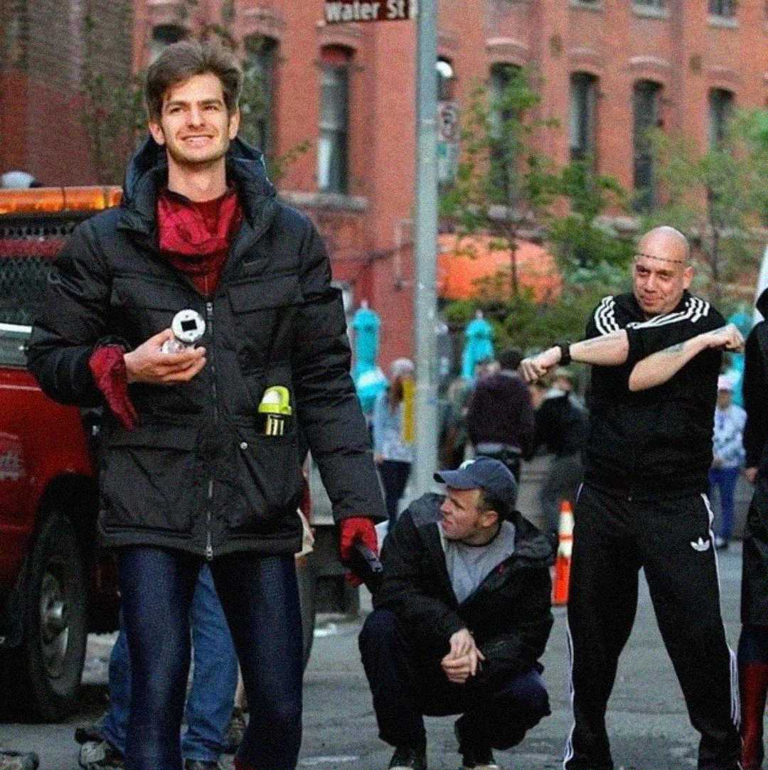 A smiling man in a puffy black jacket and shiny blue pants stands on a city street holding a camera, while two men behind him, one in black athletic wear, appear to be stretching or warming up. Brick buildings are visible in the background.