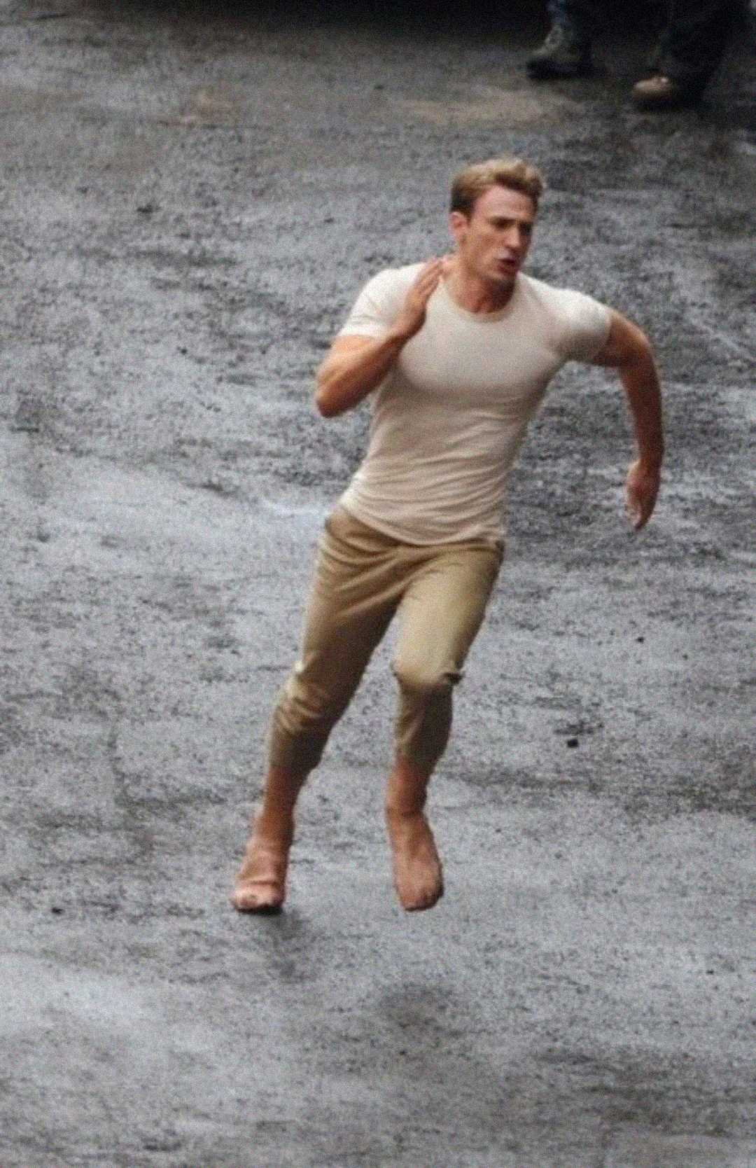 A man wearing a white T-shirt and beige pants is running barefoot on a wet, gray pavement, appearing to be in motion and focused.