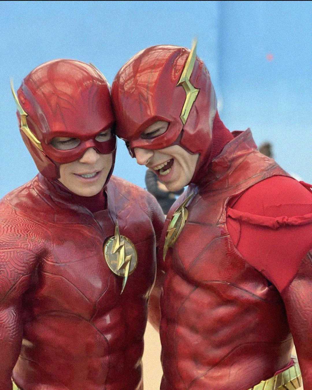 Two people dressed as The Flash, wearing red superhero costumes with gold lightning bolt emblems and matching helmets, stand close together and smile, with a blurred background.