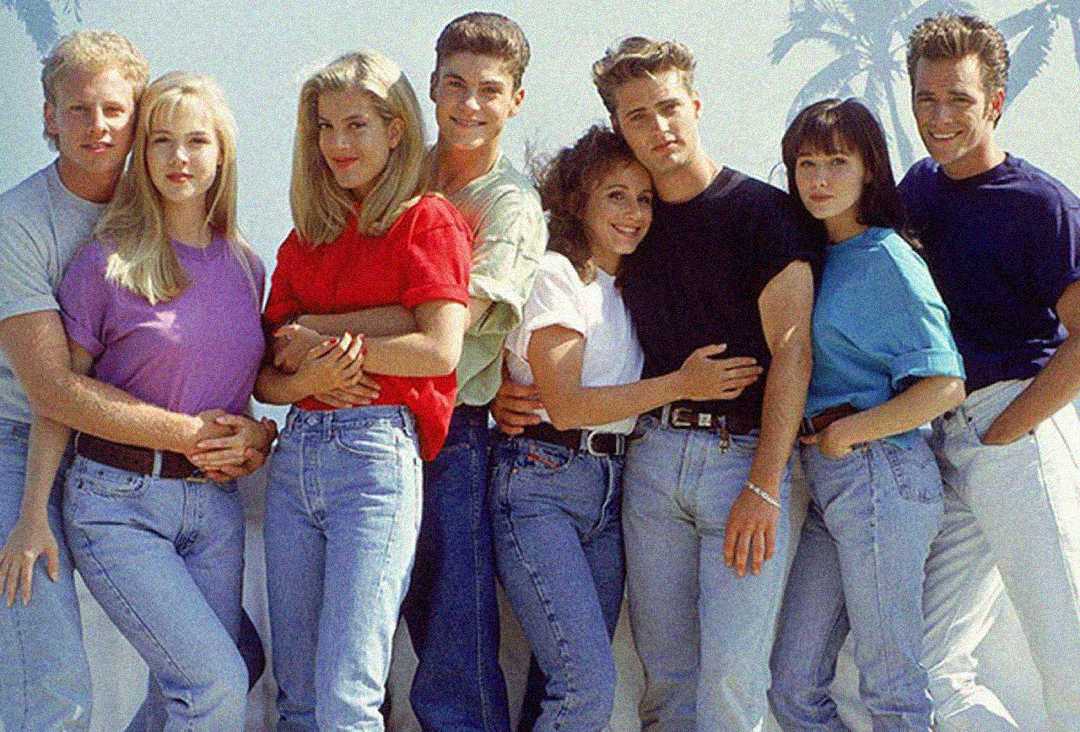 Eight young adults stand closely together, smiling and embracing, all dressed in casual 1990s clothing with light blue jeans and colorful tops. Palm trees are faintly visible in the background.