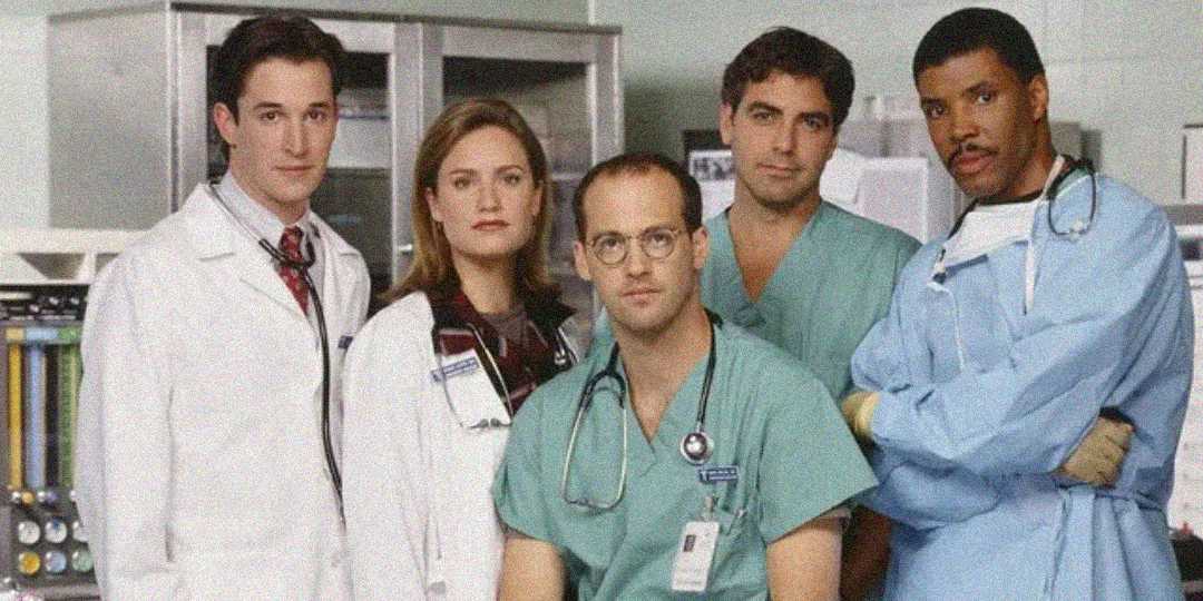 Five doctors in medical attire, including lab coats and scrubs, stand together in a hospital setting, looking seriously at the camera with medical equipment visible in the background.