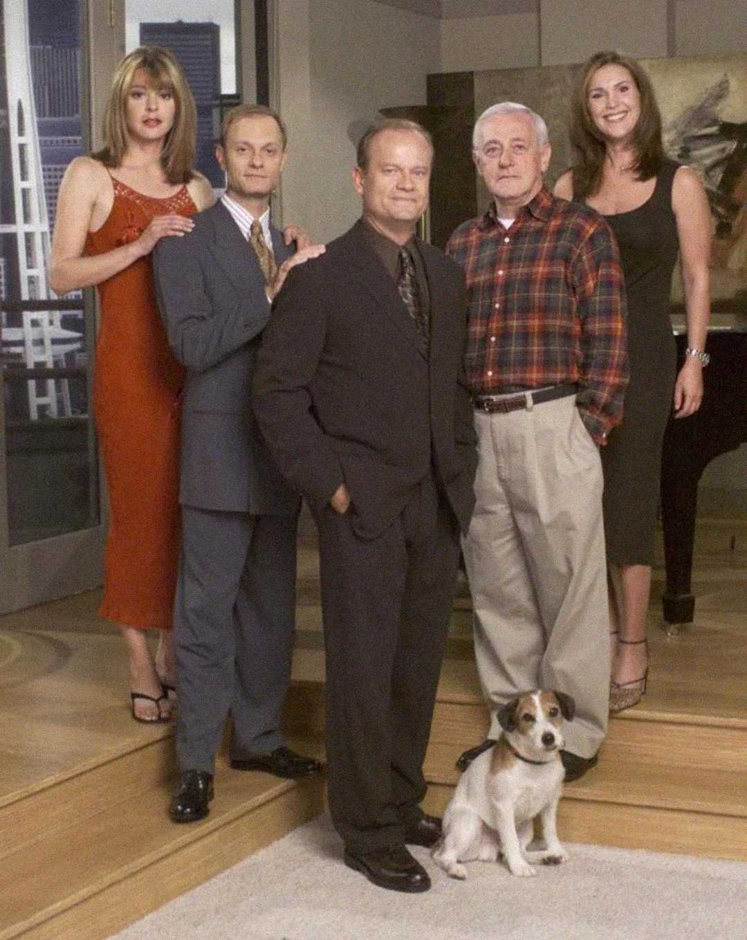 Five people and a small dog pose together inside a modern living room. Two women and three men stand, dressed in semi-formal attire, while the dog sits on the floor in front of them.
