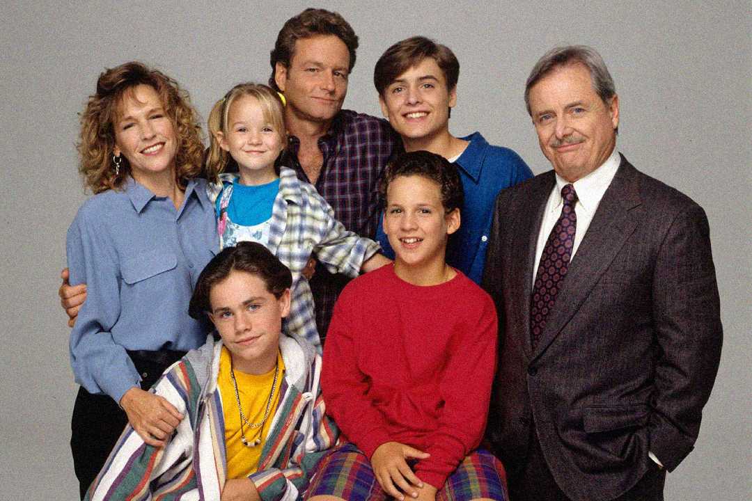 A group of seven people, consisting of four adults and three children, pose together and smile against a plain light gray background. They are dressed in casual and semi-formal 1990s-style clothing.