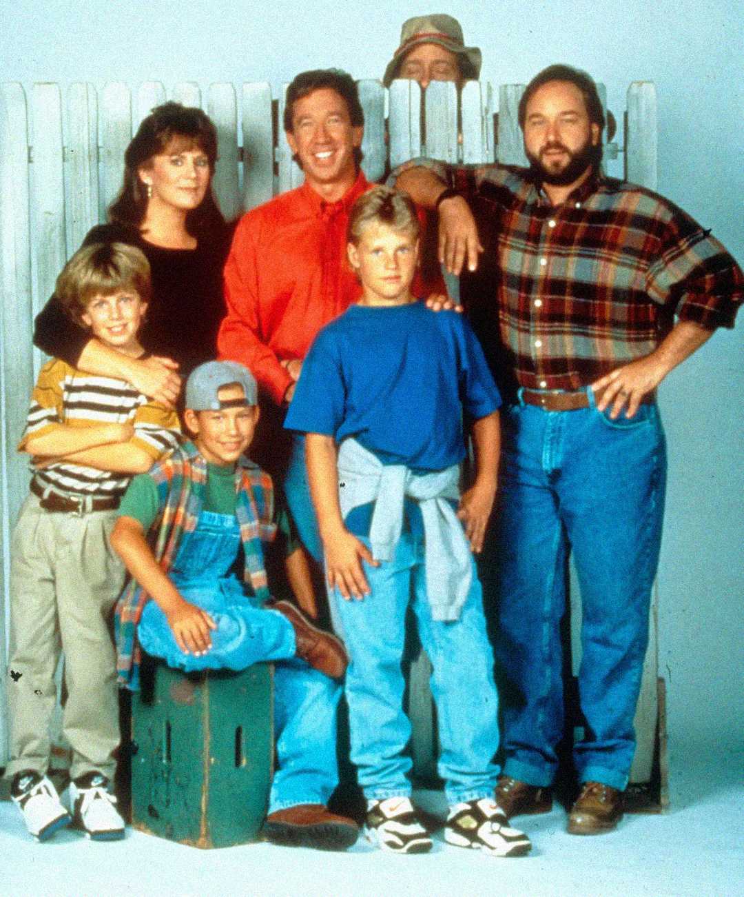 A group of seven people pose in front of a white wooden fence; three adults stand behind four boys, with one adult peeking over the fence. The group appears cheerful and casually dressed in '90s-era clothing.