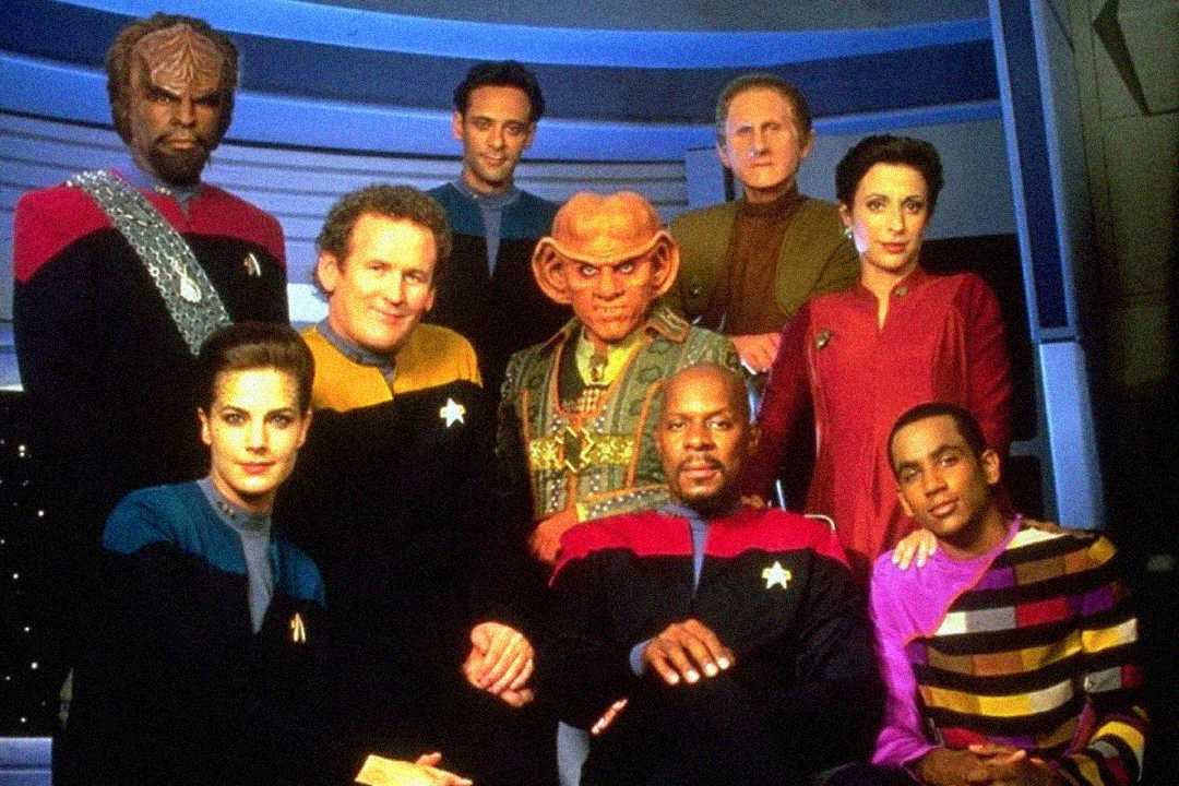 Nine people in colorful Star Trek uniforms and distinctive makeup pose together on a futuristic spaceship set, representing a diverse science fiction crew from "Star Trek: Deep Space Nine.