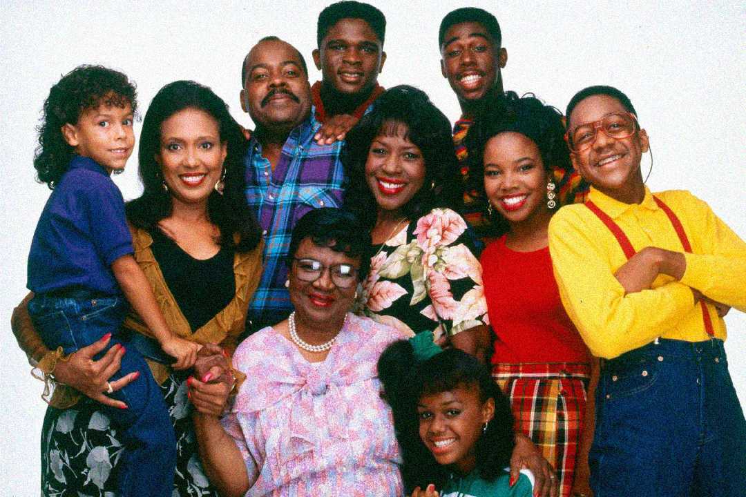 A group of eleven people, both adults and children, stand together smiling against a plain white background. They are dressed in colorful, casual 90s clothing, posing closely in a cheerful family portrait.