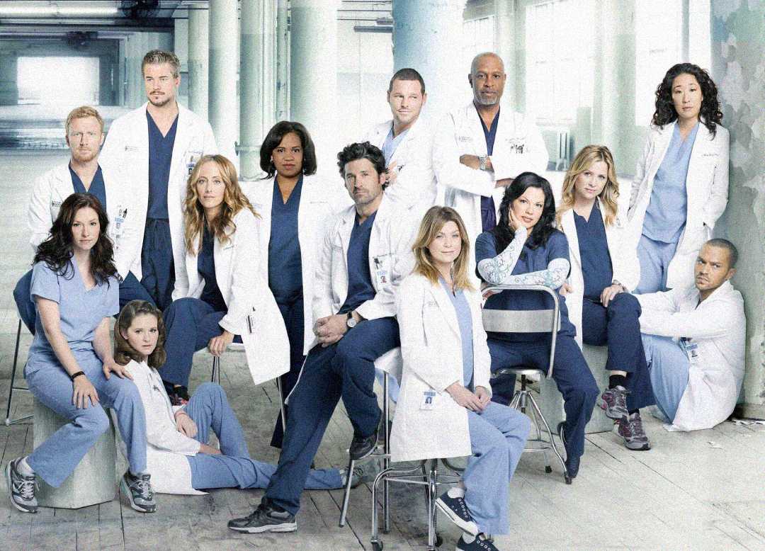 A group of medical professionals in scrubs and white coats pose together in a bright, industrial-style hallway, some standing and some seated, looking serious and confident.