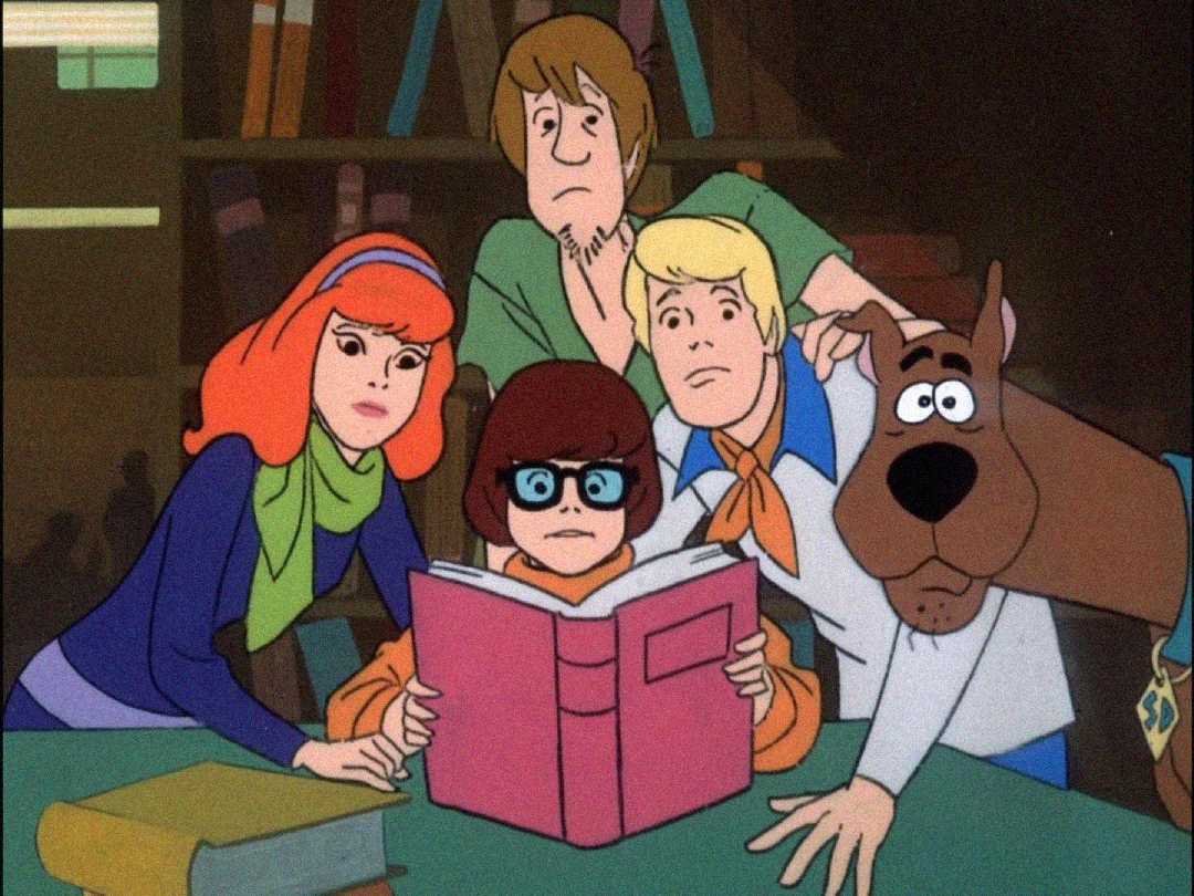 Five animated characters—Daphne, Velma, Shaggy, Fred, and Scooby-Doo—gather closely around an open pink book, looking surprised and curious in a dimly lit library setting.