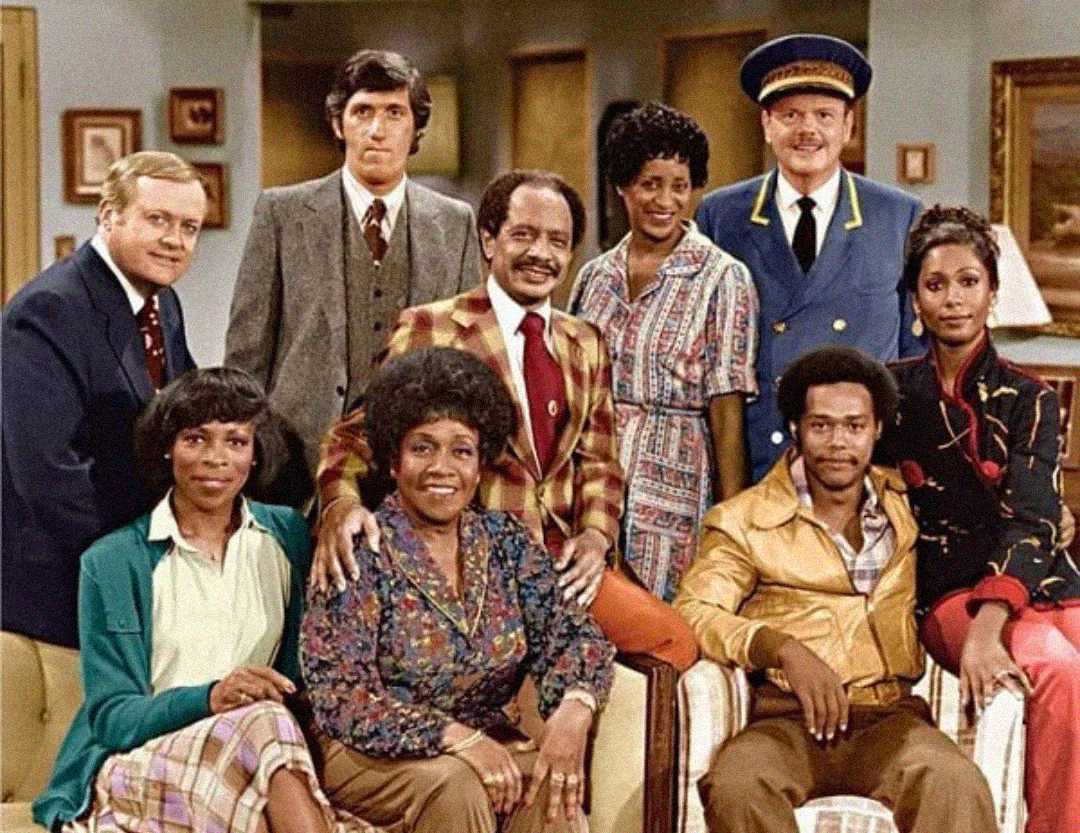 A group of nine people, dressed in 1970s-style clothing, pose and smile together in a living room setting. Some are seated while others stand behind them; one man wears a blue doorman's uniform and cap.