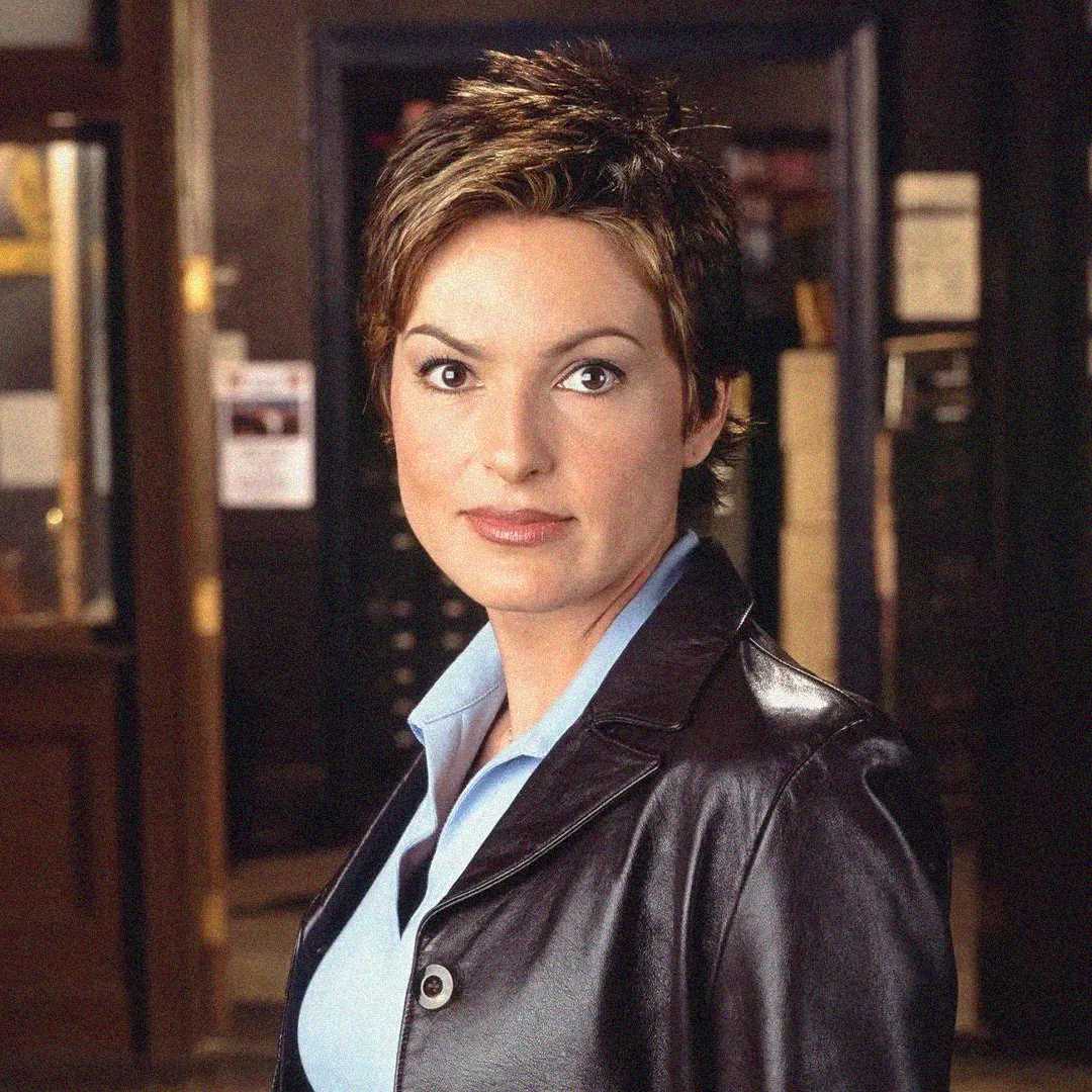 A woman with short brown hair wearing a black leather jacket over a light blue shirt stands indoors, looking directly at the camera with a neutral expression. The background is slightly blurred.