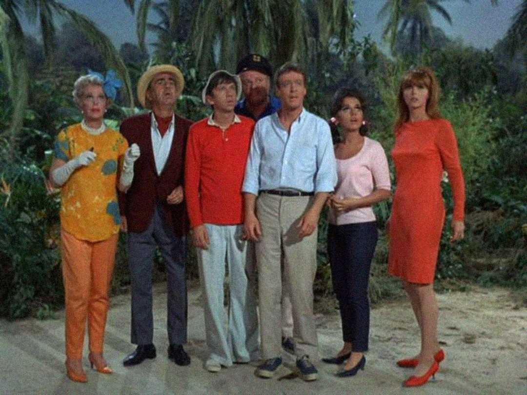 Six people stand together outdoors in tropical surroundings, all looking upward with surprised or concerned expressions. Palm trees and greenery are visible in the background.