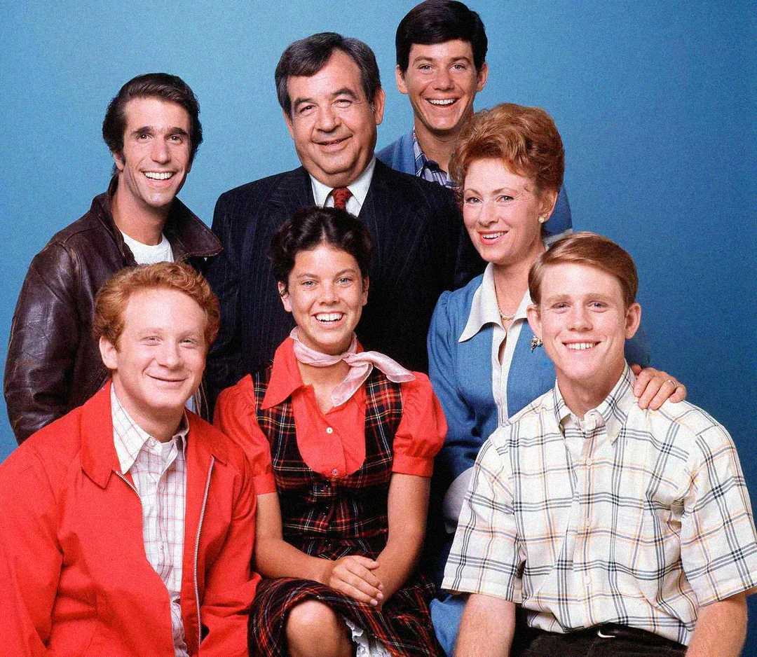 A group of seven people, four men and three women, are smiling and posing together in front of a blue background. They are dressed in casual and semi-formal 1950s-style clothing.
