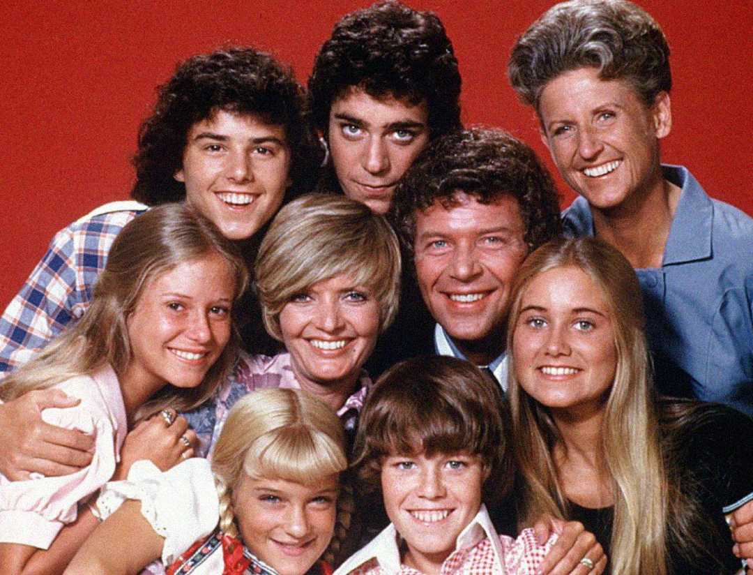 A group of nine people, including adults and children, pose closely together and smile in front of a red background. They appear cheerful and friendly, with their heads close and arms around each other.
