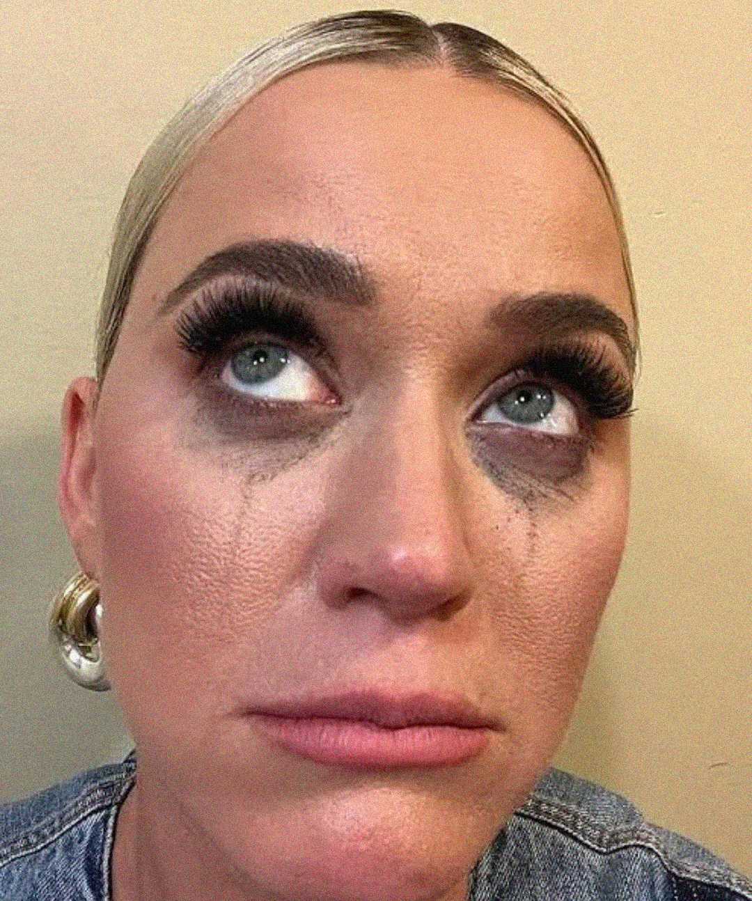 A woman with platinum blonde hair and large earrings looks upward with a sad expression. Her mascara is smudged beneath her eyes, suggesting she has been crying. She is wearing a denim top and heavy makeup.