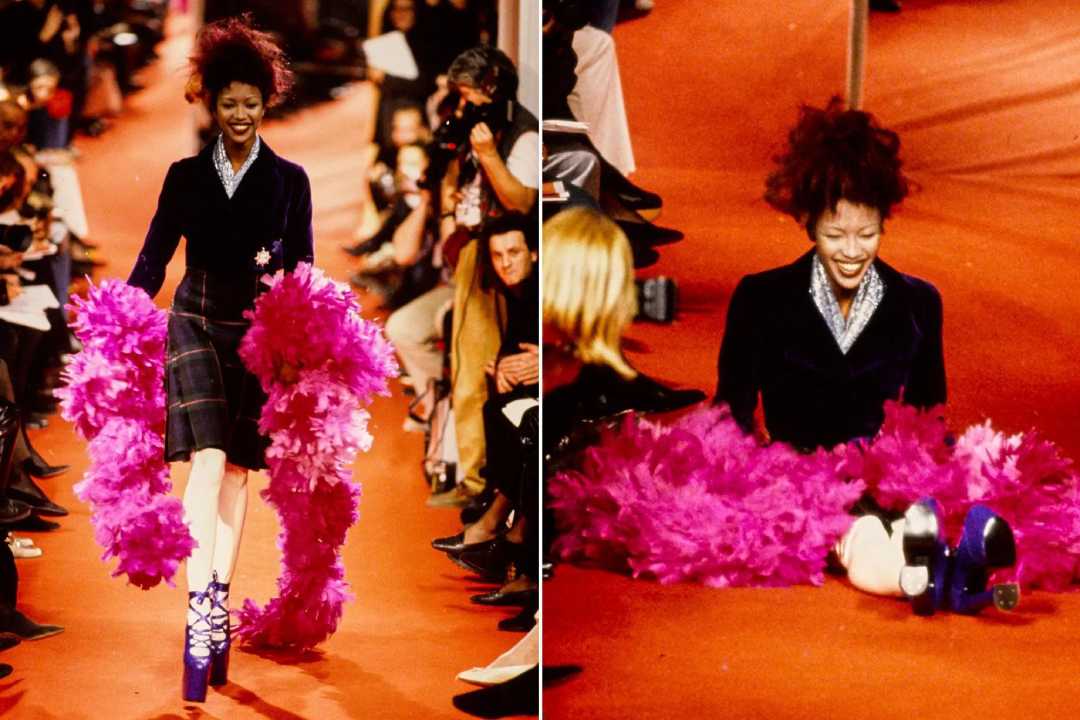 A model in a dark outfit with bright pink feathered accessories walks down a red runway and then falls, smiling as she sits on the floor while the audience watches.