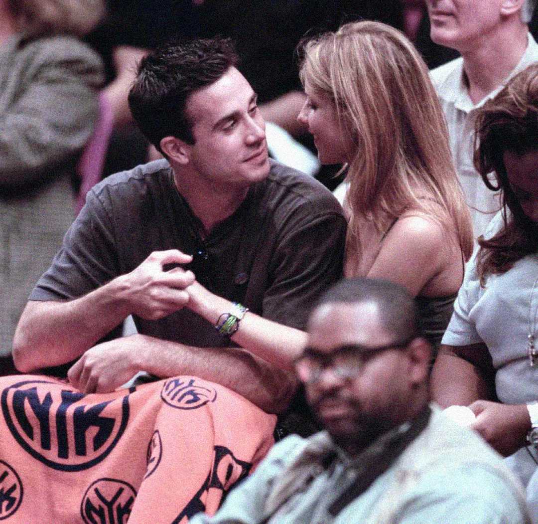 A man and woman sit closely together in a crowd at a sports event, smiling and holding hands, with an orange Knicks blanket draped over their laps. People are seated around them, one in the foreground wearing glasses.