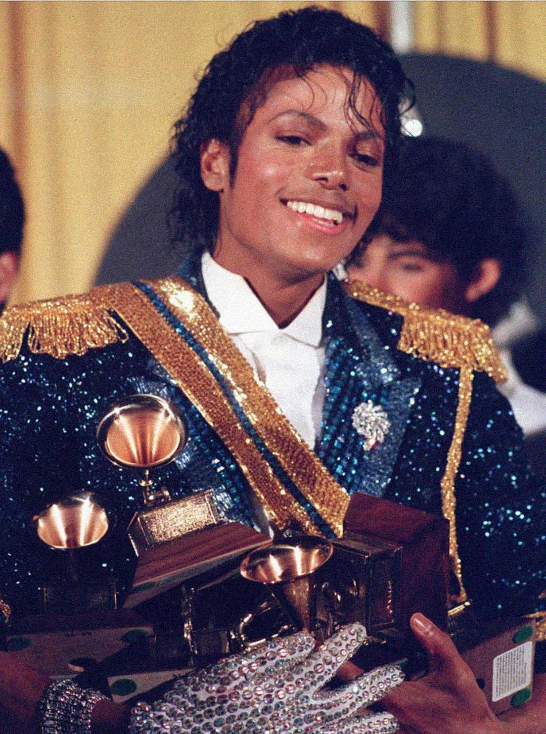 A man wearing a sequined jacket and glove smiles while holding several Grammy Awards, with a yellow curtain in the background.
