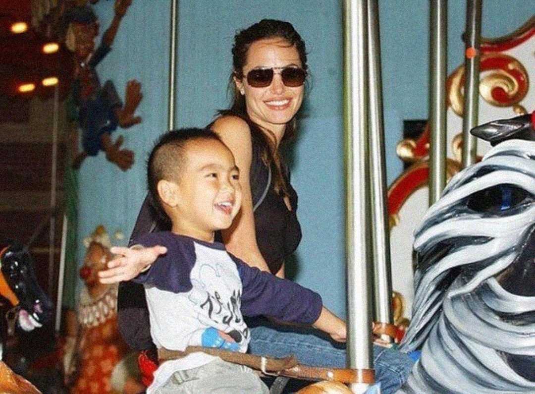 A woman wearing sunglasses and a black top smiles while riding a carousel with a cheerful young boy who has his arms outstretched. They are sitting on a zebra carousel animal, and colorful decorations are in the background.