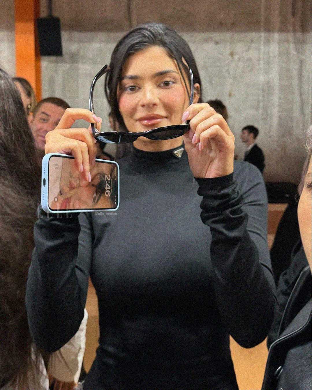 A woman with long dark hair wearing a black top holds a pair of sunglasses and a phone, smiling at the camera in a crowded indoor setting.