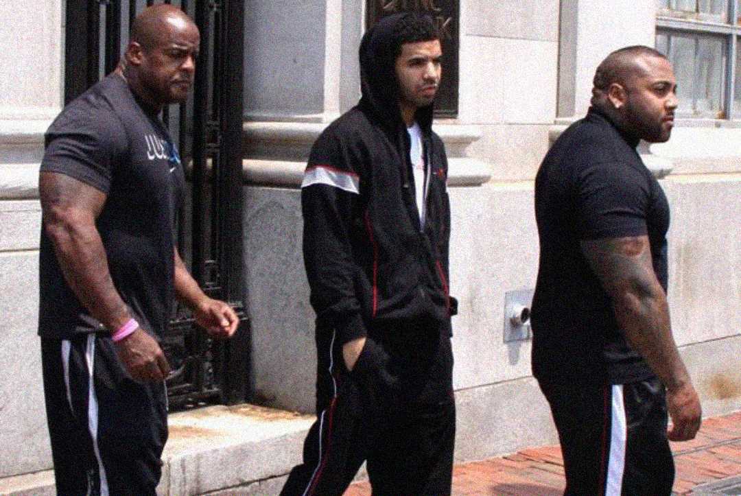 Drake walks between two large men in black athletic wear. One man on the left has a pink bracelet and the other on the right has tattoos on his arm.