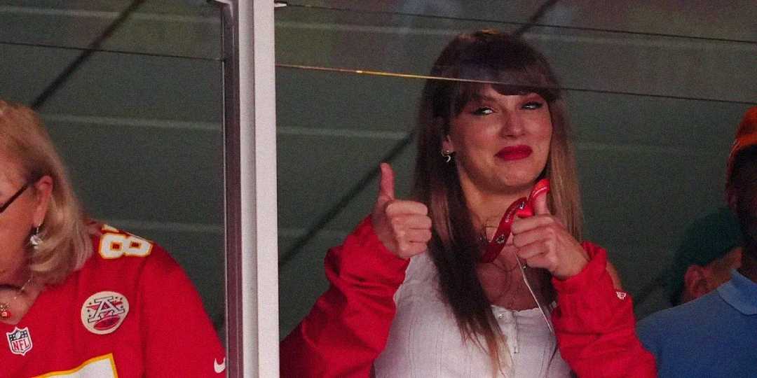 A woman with long brown hair and red lipstick smiles and gives two thumbs up while standing indoors. She is wearing a red jacket and holding a lanyard. Others in her suite appear to be watching a sports event.