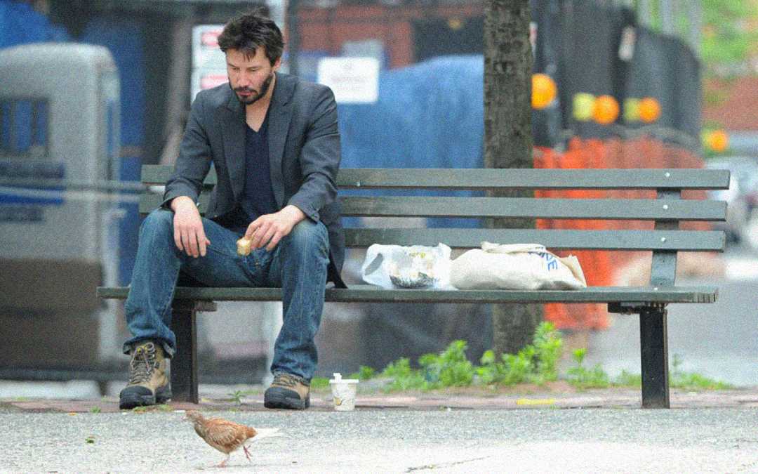 A man in casual clothes sits alone on a bench, looking down while holding food. There are bags and a drink beside him on the bench, and a bird stands on the ground in front of him. The background is urban and blurred.