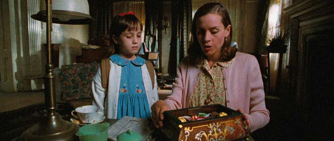A young girl in a blue dress stands beside a woman in a pink cardigan, who is opening a tin box filled with chocolates at a table in a cozy, dimly lit room.