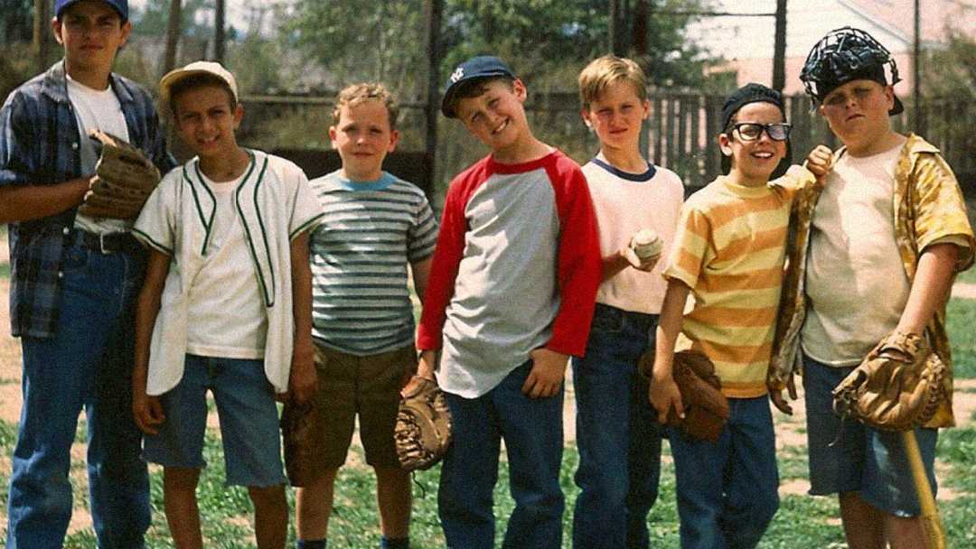 Seven boys wearing casual summer clothes and baseball gloves stand together on a baseball field, smiling and posing for the camera. A fence and trees are visible in the background.