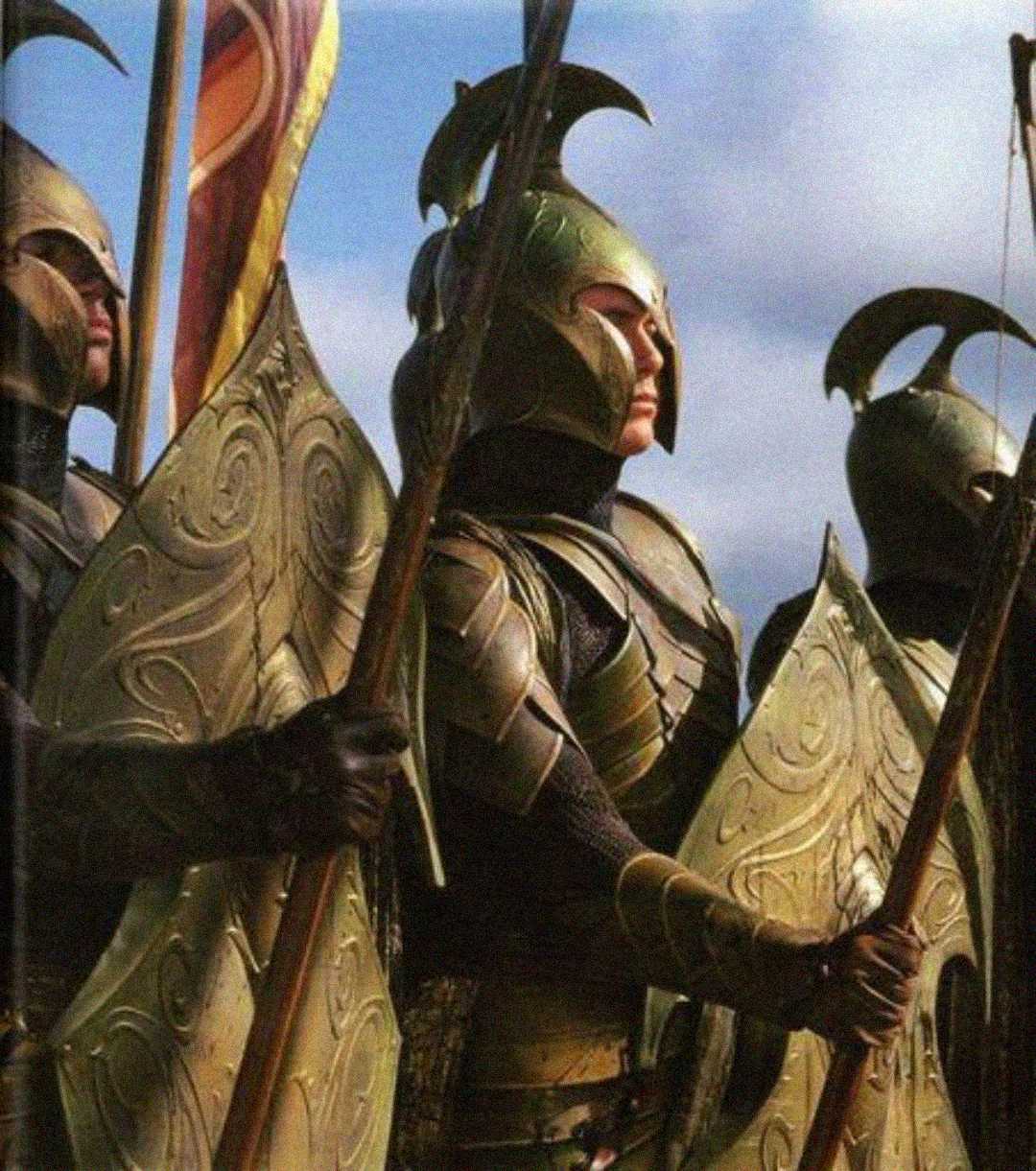 A group of armored warriors wearing ornate gold helmets and holding large, decorated shields and spears stand in formation under a blue sky.