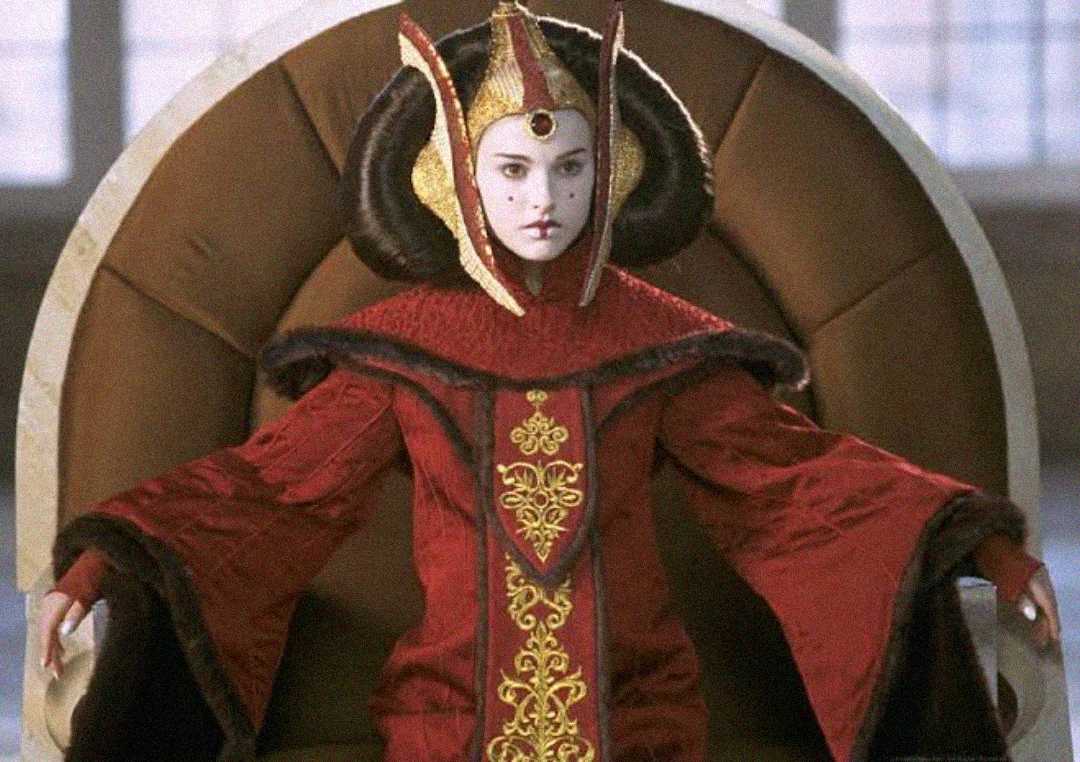 A woman in elaborate red and gold royal attire with a tall, ornate headdress sits on a large, round throne, her face painted white with red accents, exuding a regal and serious expression.