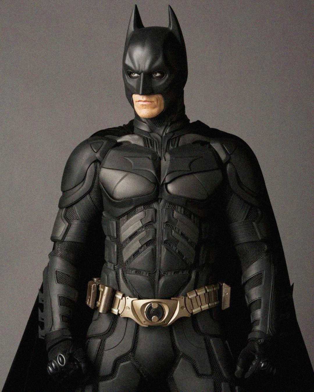 A person dressed as Batman stands facing forward, wearing a detailed black armored suit with a gold utility belt and a black cape against a plain, neutral background.