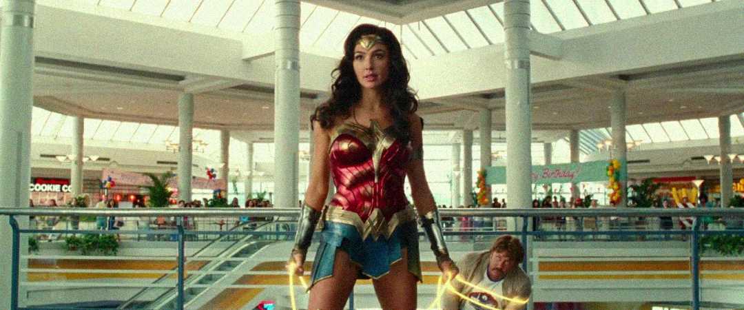 A woman in a red and gold superhero costume stands confidently in a brightly lit shopping mall, holding a glowing golden lasso. People and shops are visible in the background.