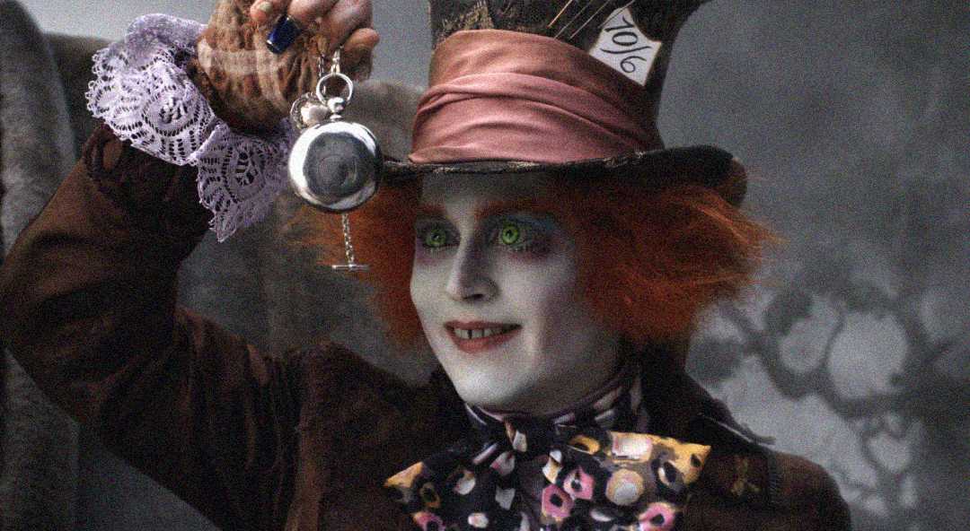A person dressed as the Mad Hatter with bright orange hair, a top hat with a "10/6" card, pale face makeup, colorful clothes, and holding a pocket watch up in one hand.