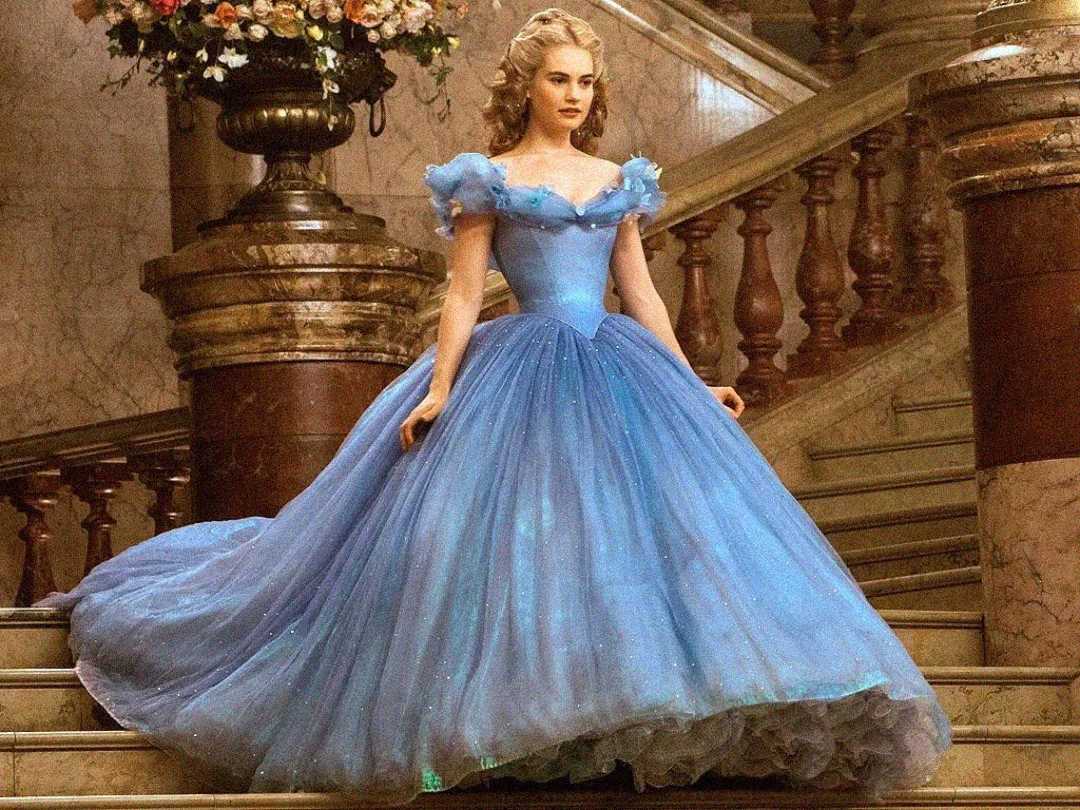 A woman in a sparkling blue ball gown with puffed sleeves stands gracefully on a grand marble staircase, with a large floral arrangement visible behind her.