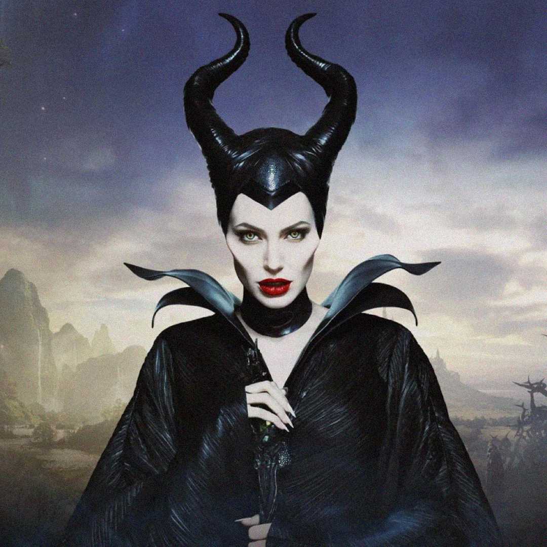 A woman dressed as Maleficent wears a black horned headdress, dramatic makeup with red lips, and a black high-collared cape, standing against a misty, mysterious landscape.