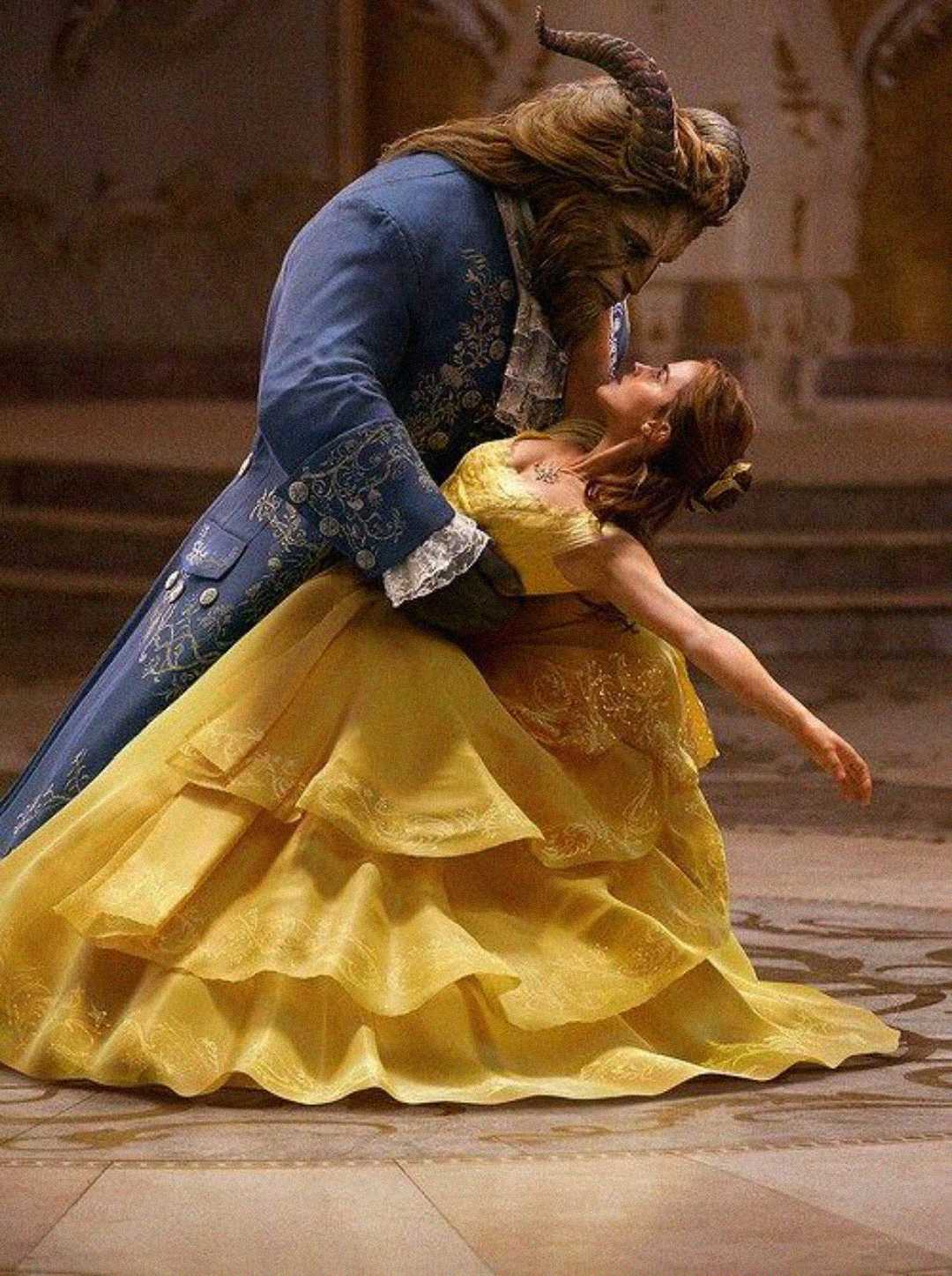A large, beastly figure in an ornate blue coat dances with a woman in a flowing yellow gown. They are gazing at each other, mid-dance, in an elegant, softly lit ballroom.