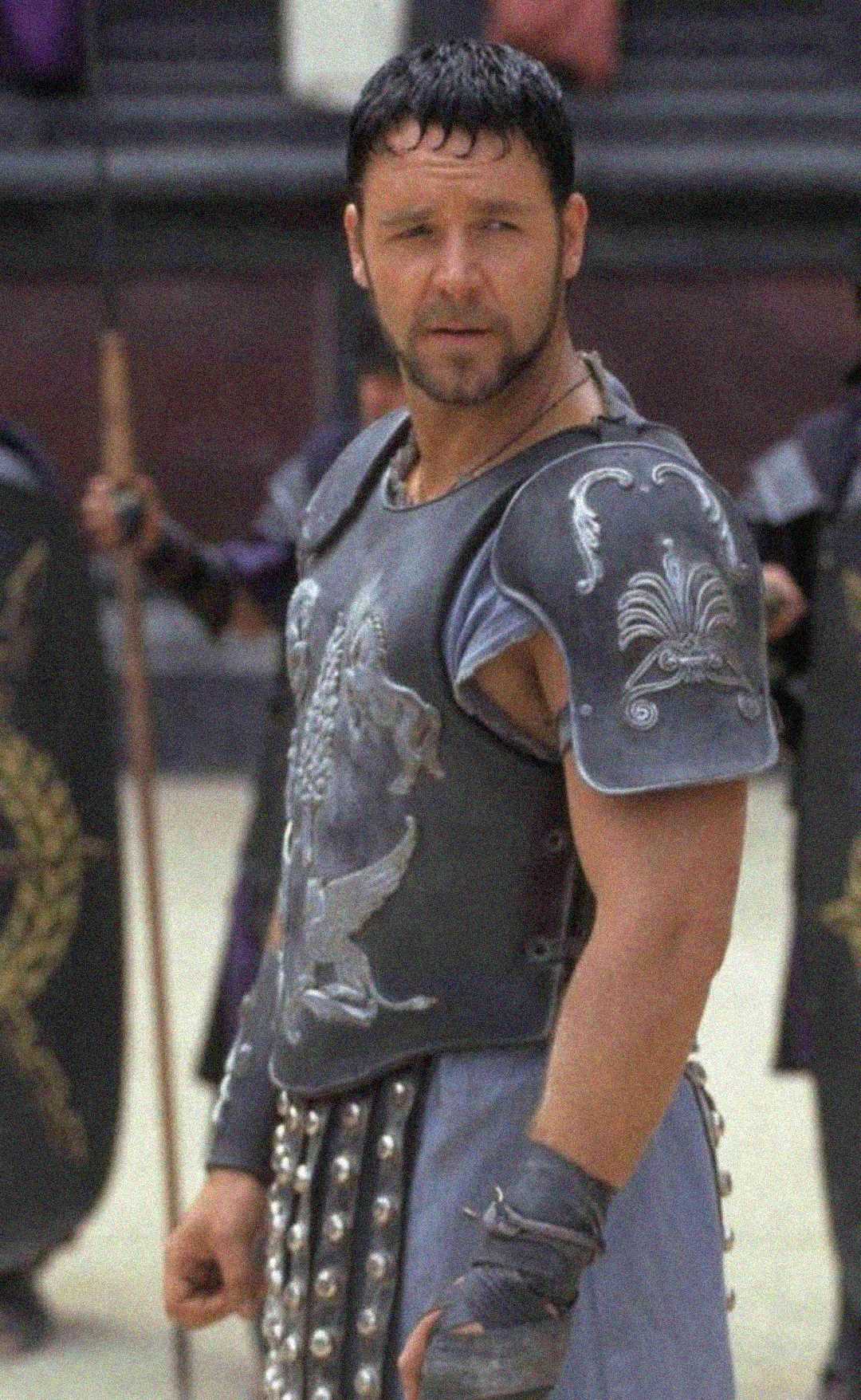 A man in ancient Roman-style armor stands in an arena, looking to the side. He has short dark hair and is surrounded by soldiers holding shields in the background.