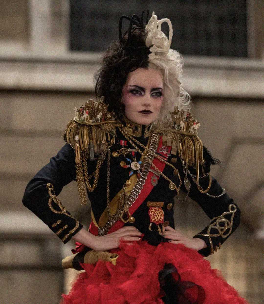 A person in dramatic makeup and costume poses confidently. They wear a black and gold military-style jacket with ornate decorations, red ribbons, and gold fringe, paired with a voluminous red and black skirt. Their hair is black and white.