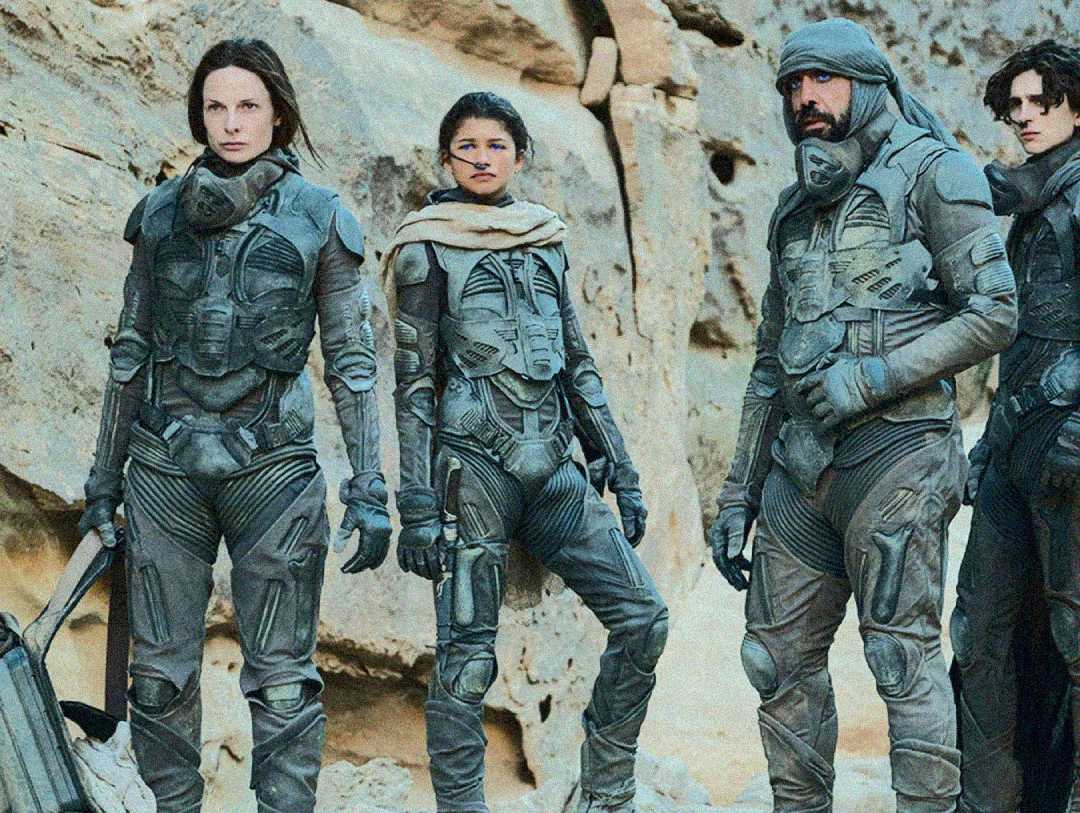 Four people in futuristic armor stand together in a rocky desert landscape, looking serious and alert. They wear gray suits with high collars and various gear, blending into the sandy environment.