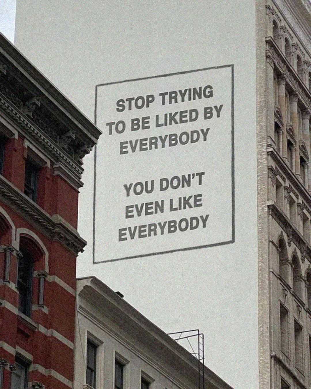 A large mural on the side of a city building reads, "STOP TRYING TO BE LIKED BY EVERYBODY. YOU DON'T EVEN LIKE EVERYBODY." The surrounding buildings are older, with decorative brick and stone facades.