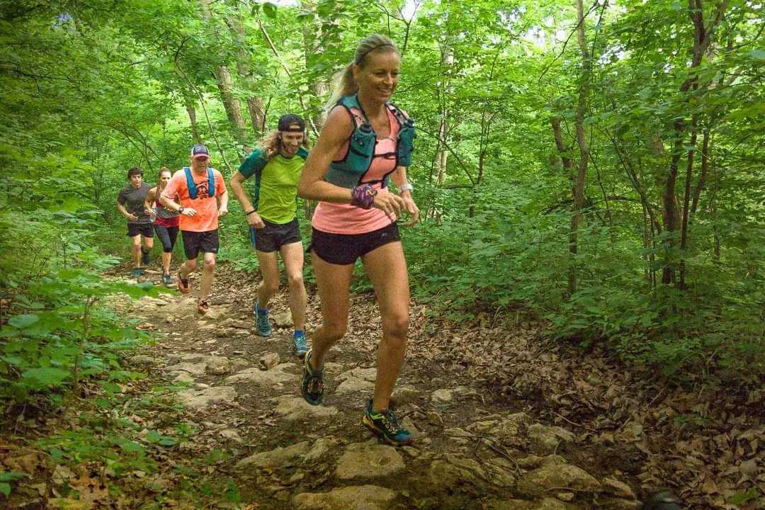 A group of people run on a rocky trail through a lush, green forest. The leading runner, wearing a pink top and hydration vest, smiles as others follow closely behind. Leaves and trees surround the path.