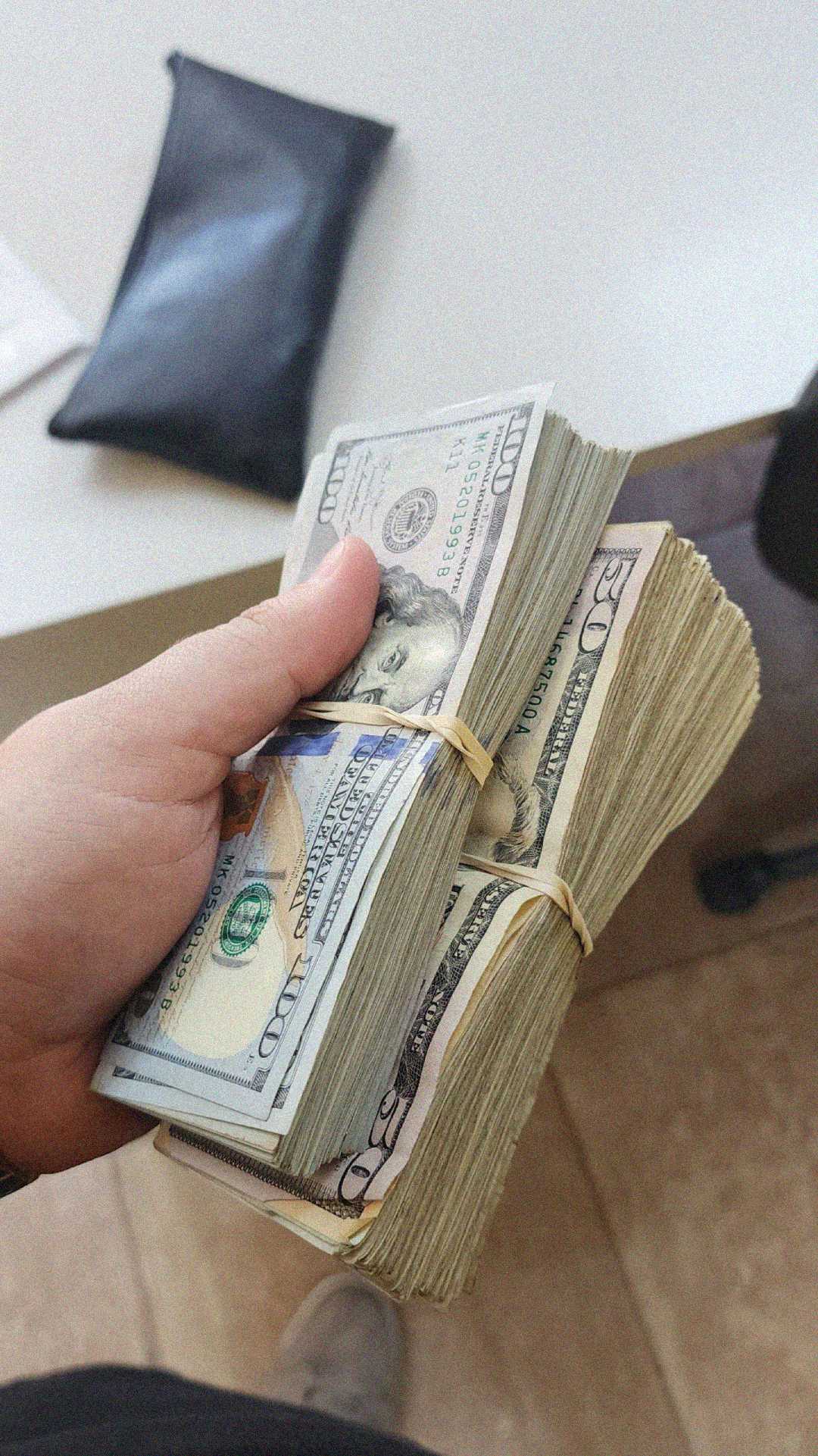 A hand holding two thick bundles of U.S. dollar bills secured with rubber bands, with a black pouch and a white surface visible in the background.