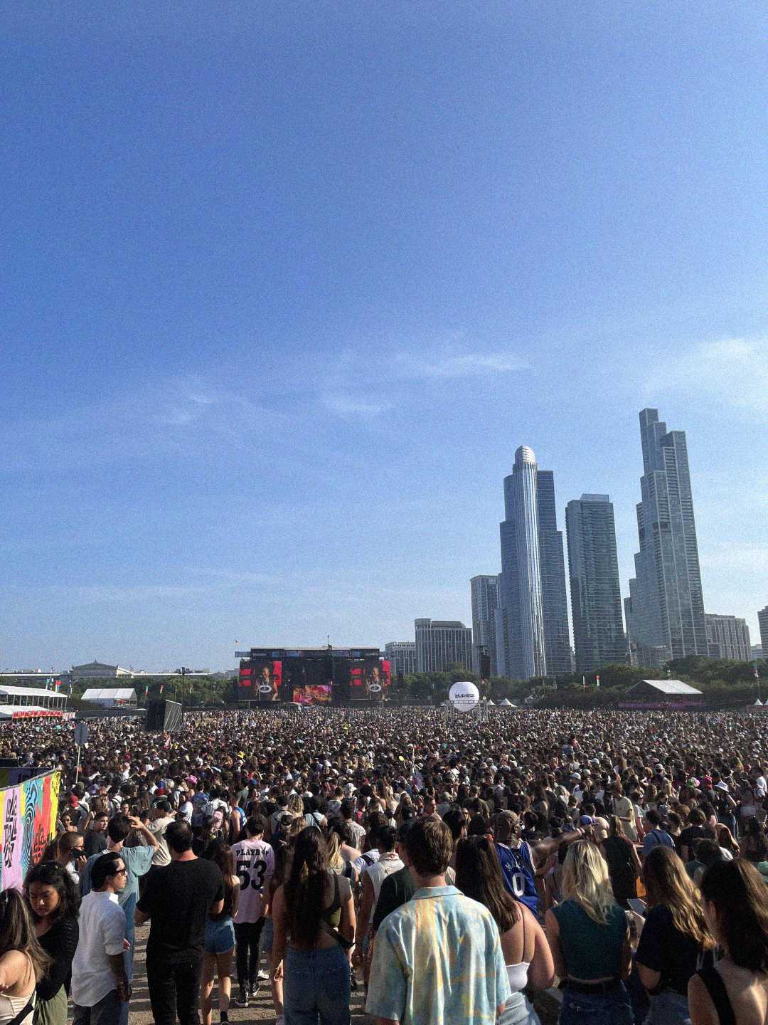 A large crowd attends an outdoor music festival in a city park, with tall skyscrapers in the background under a clear blue sky. People face a distant stage with large screens.