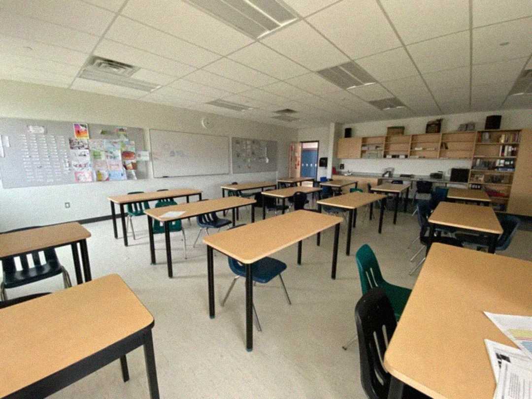An empty classroom with wooden desks and plastic chairs arranged in rows. The room has whiteboards, posters on the wall, shelves with books and supplies, and bright overhead lighting.